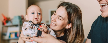 En mamma håller upp sin bäbis, två armar till från en andra förälder syns i förgrunden.