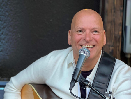 En leende man spelar akustisk gitarr och sjunger i mikrofon, klädd i vit tröja.