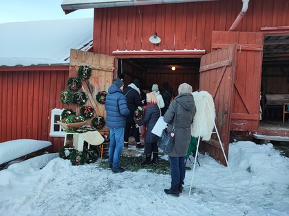 Personer i vinterkläder står i kö utanför en röd träbyggnad med julkransar och snö på marken.