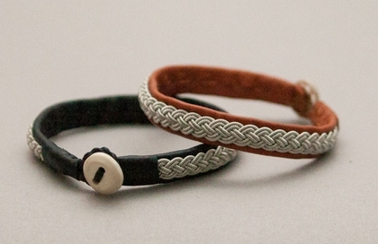 Två armband, ett svart och ett brunt, med flätade silverfärgade detaljer, ligger på ett ljust underlag.