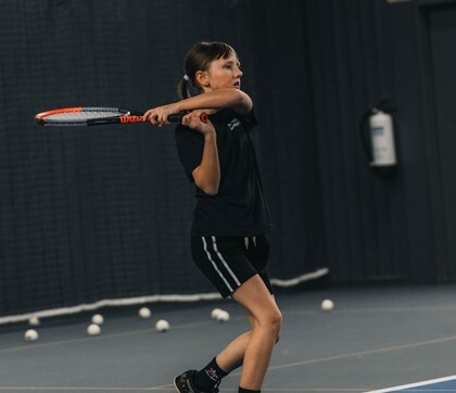 En person spelar tennis inomhus och svingar ett racket på en tennisbana med flera bollar på golvet.