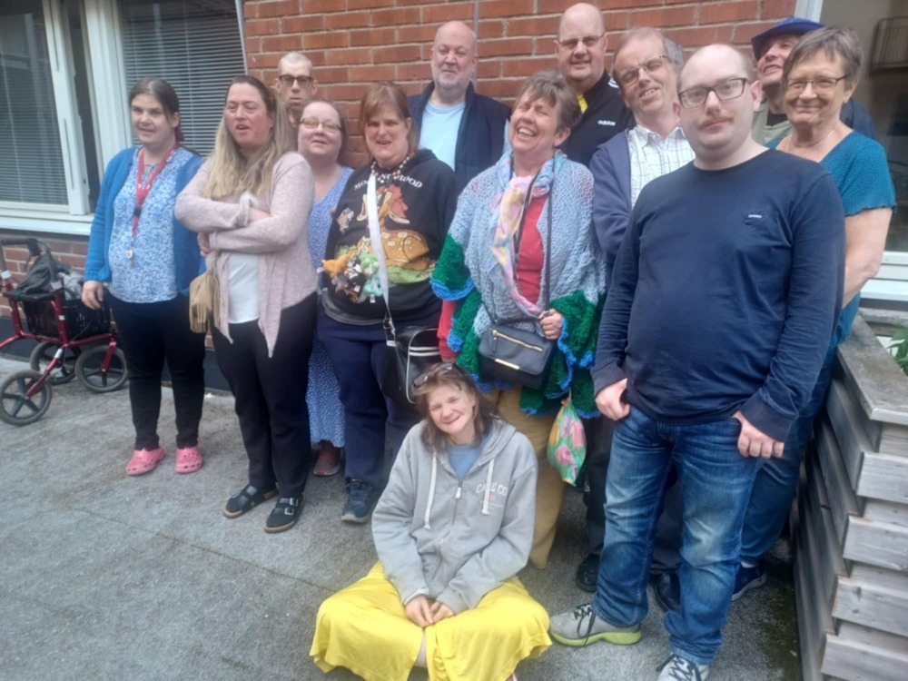 En grupp människor står och sitter utomhus framför en tegelvägg, några med färgglada kläder.