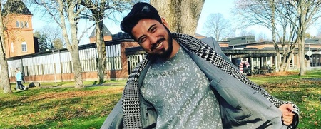 En leende man står utomhus i en park, håller upp sin jacka och poserar mot kameran.