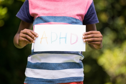 Ett barn håller upp en skylt med texten 'ADHD' skrivet i färgglada bokstäver utomhus.
