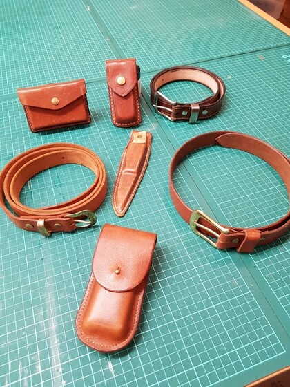 Flera handgjorda läderprodukter, inklusive bälten, fodral och armband, ligger på ett grönt skärmatta.