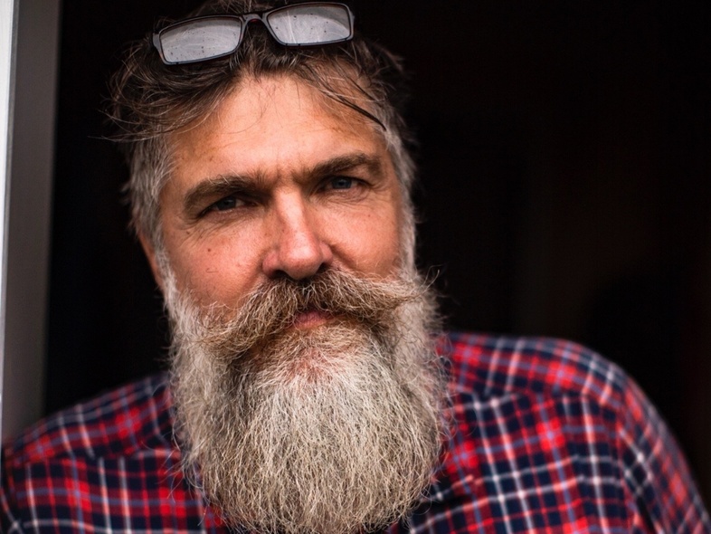 Man med stort grått skägg och glasögon på huvudet, bär rödblå rutig skjorta, står mot mörk bakgrund.