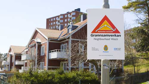 Skylt för grannsamverkan sitter på stolpe framför röda trähus och ett höghus i bakgrunden.