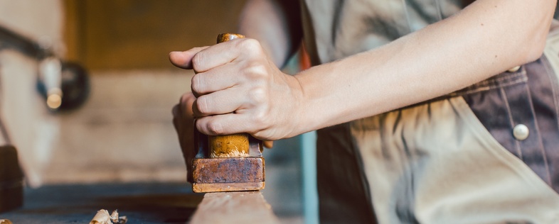 En person i arbetskläder hyvlar trä i en snickarverkstad, fokus på händer och verktyg.