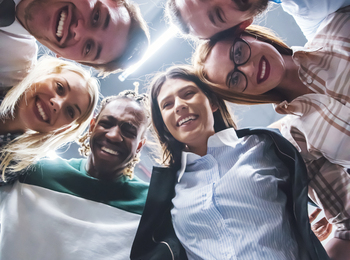 En grupp glada människor står i en cirkel och tittar ner mot kameran, fotograferade underifrån.