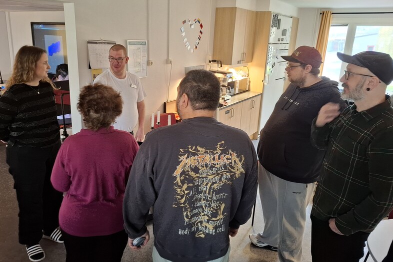 En grupp människor står i en cirkel och samtalar i ett ljust rum med köksinredning.