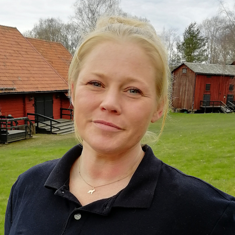 Kvinna med blont hår står utomhus framför röda trähus och gräsmatta, bär mörkblå pikétröja.