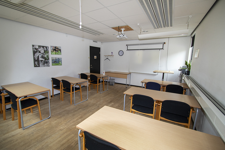 Ett tomt klassrum med flera bord och stolar, vita väggar och en projektorduk framtill.
