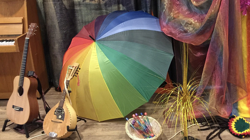En regnbågsfärgad paraply står uppfällt bredvid en gitarr, mandolin, penslar och färgglada tyger.
