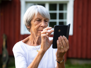 Äldre kvinna med grått hår använder en smartphone utomhus framför ett rött trähus.