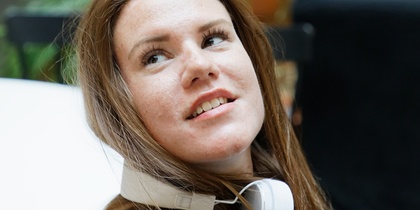 Kvinna med långt brunt hår och hörlurar runt halsen sitter inomhus och tittar uppåt.