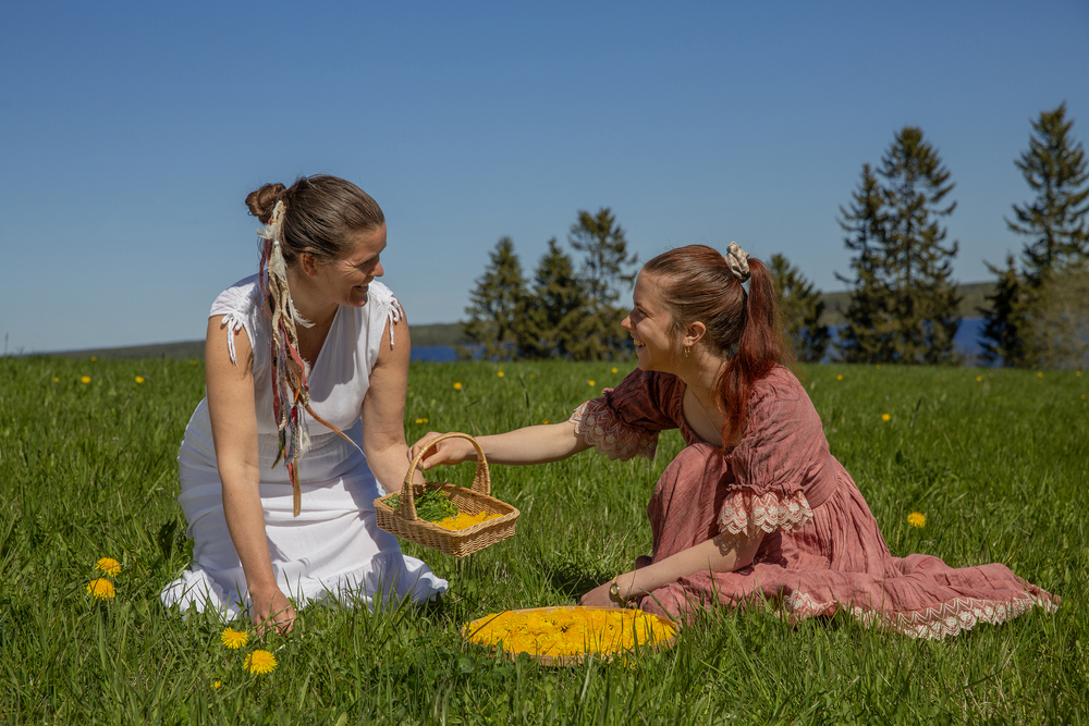 Två kvinnor i klänningar sitter på en grön äng och plockar blommor i en korg under en klarblå himmel.