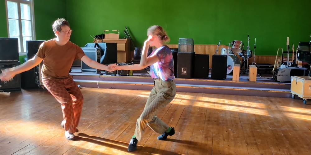 Två personer dansar i ett solbelyst rum med trägolv och musikutrustning i bakgrunden.