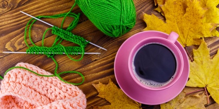 Stickning med grönt garn, rosa stickat plagg, rosa kaffekopp och gula höstlöv på ett träbord.