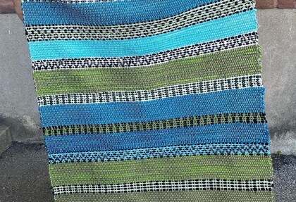 En handvävd matta med ränder i blått, grönt, svart och vitt står lutad mot en vägg utomhus.