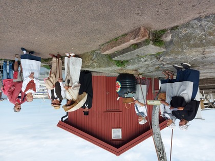 En grupp människor i historiska kläder står utomhus vid en röd träbyggnad, några samtalar och andra tittar på.