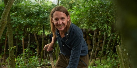 Person arbetar med en grep i en kompostbehållare i en grönskande trädgård.