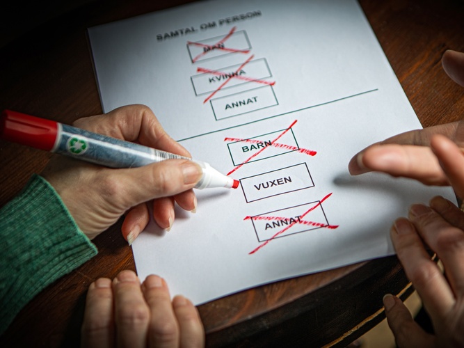 Två händer markerar och kryssar i rutor på ett formulär med en röd penna.