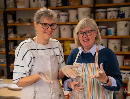 Två leende kvinnor med glasögon och förkläden står i en keramikverkstad med händerna täckta av lera.