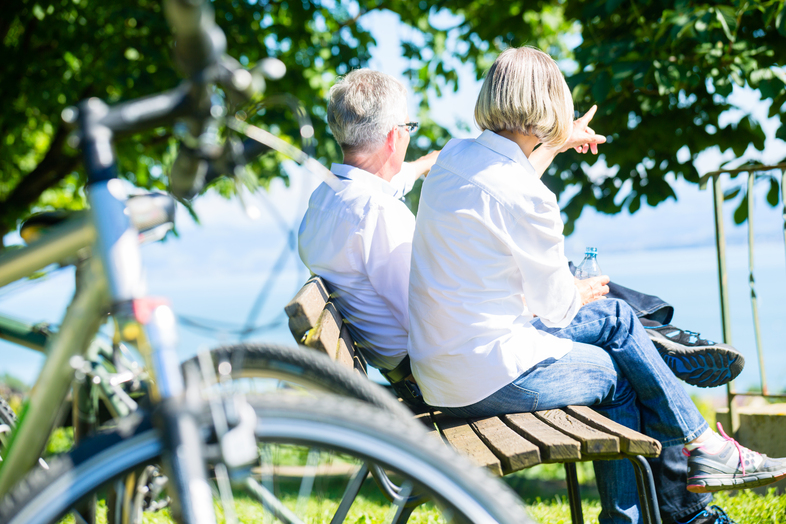 Två äldre personer sitter på en bänk vid vattnet, omgivna av cyklar och grönska, och pekar mot utsikten.