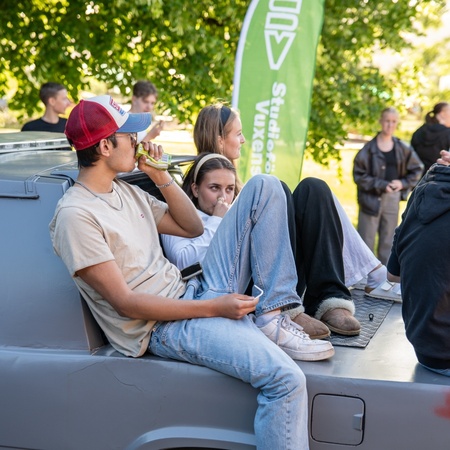 Två unga personer sitter avslappnat på flaket av en bil under ett utomhusevenemang.
