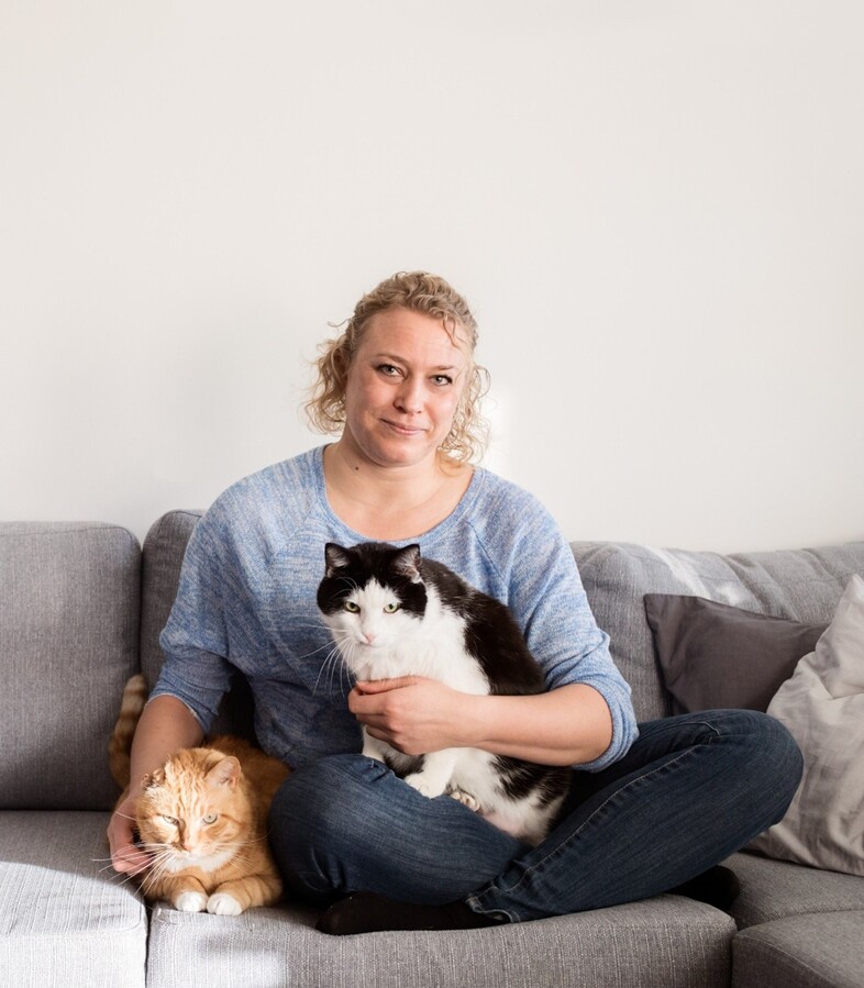 Kvinna sitter på en soffa med en svartvit katt i famnen och en orange katt bredvid sig.