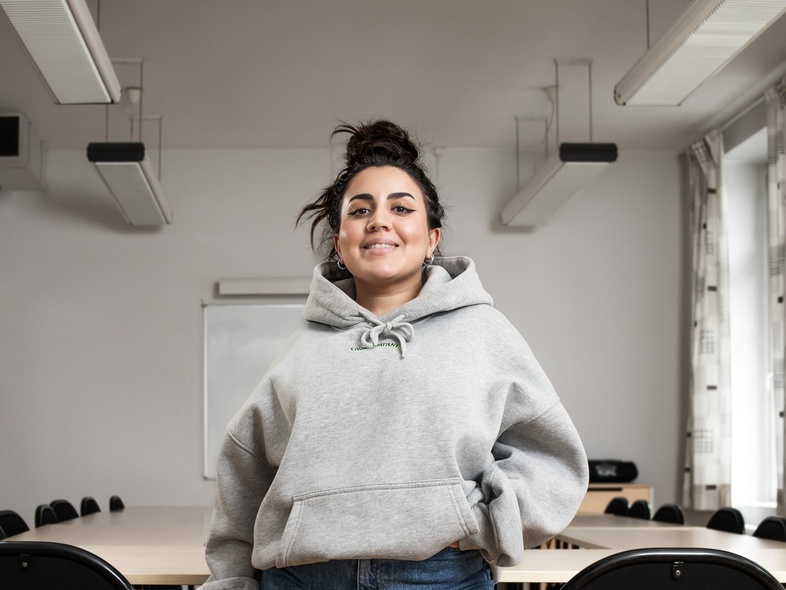 Ung kvinna i grå huvtröja står leende i ett tomt klassrum med bord och stolar.