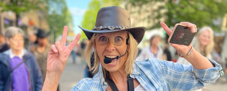 Kvinna med cowboyhatt och mikrofon gör segertecken på en stadsgata, leende och energisk.