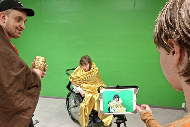 Tre personer spelar in en film framför en grön vägg, en sitter i rullstol med gul mantel, en filmar med surfplatta.
