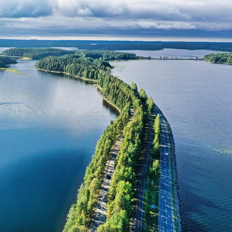Lång smal väg med träd på båda sidor som går genom en stor sjö under molnig himmel.