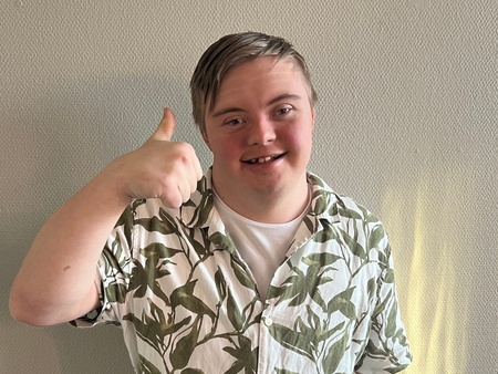 Ung man med blont hår ger tummen upp och ler, står mot en ljus vägg i solljus.