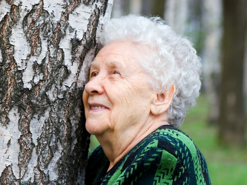 Äldre kvinna med vitt hår lutar sig mot ett träd i en skog och ler.