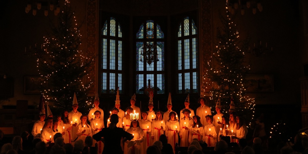 En grupp människor i vita kläder håller ljus i en mörk kyrka, omgivna av julgranar med ljusslingor.