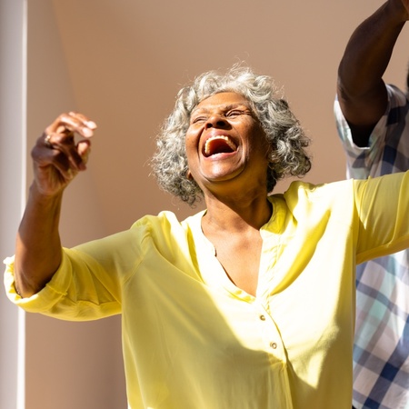 Äldre kvinna i gul skjorta skrattar och dansar med en man i ett soligt rum.