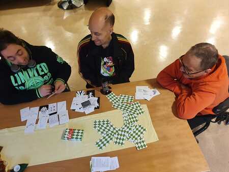 Tre personer sitter vid ett bord och spelar ett kort- eller brädspel med papper och kort.