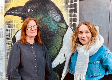 Två kvinnor står framför en väggmålning av en fågel, båda tittar mot kameran.