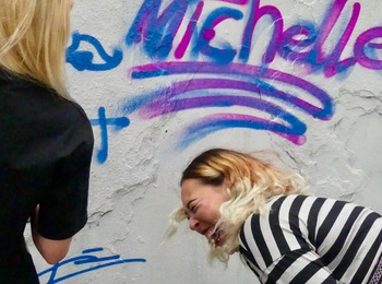 Två personer står framför Graffitiväggen i Tybbleparken. En av dem skrattar, den andra står med ryggen mot kameran. Väggen är vit, med text i rosa och lila, det står michelle.