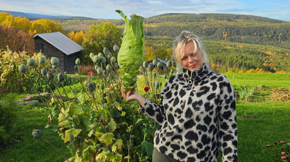 Kvinna i svartvit tröja står i trädgård och håller ett stort salladshuvud, med skog och berg i bakgrunden.