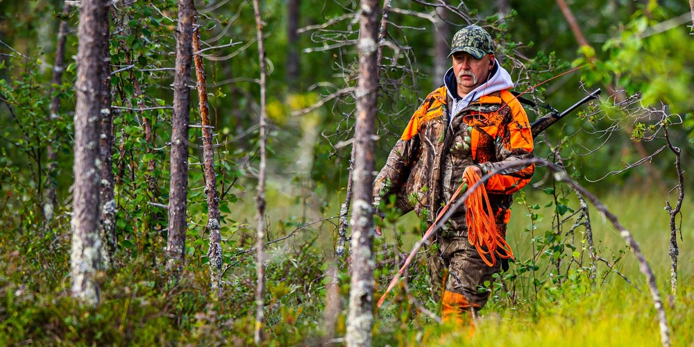 En jägare i orange och kamouflagekläder går med gevär genom en grön skog.