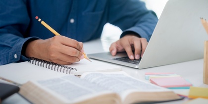 En person skriver i en anteckningsbok vid ett skrivbord med en laptop och en uppslagen bok.