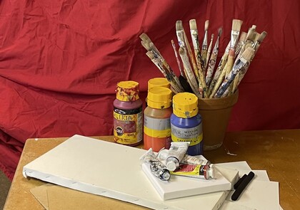 Målarpenslar i kruka, akrylfärger, tuber och små dukar på ett bord med röd bakgrund.