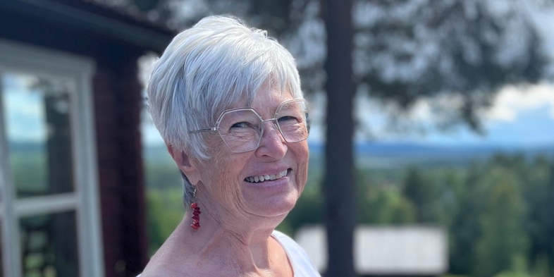 Lena Söderlund står utomhus i solsken, bär vit klänning och röda örhängen, ler mot kameran.