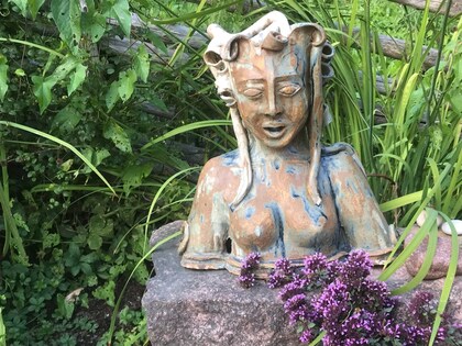 Keramikskulptur av en kvinna med långt hår står på en sten omgiven av gröna växter och lila blommor.