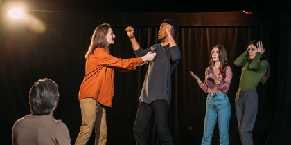 Fyra personer står på en scen och spelar teater, medan en person sitter och tittar på dem.