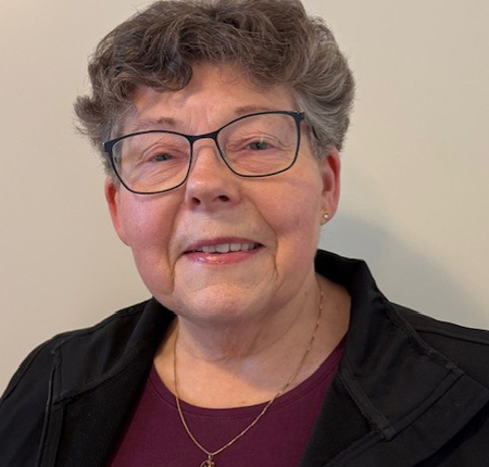 Lena Öhman har glasögon, kort grått hår och ler, bär mörk jacka och lila tröja.