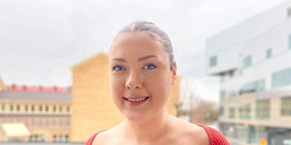 Kvinna med uppsatt hår och röd tröja står utomhus på en balkong med stadsmiljö i bakgrunden.